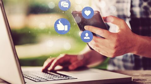 Hände tippen auf Tastatur, während Social Media Icons über Smartphone schweben