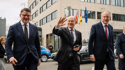 Ministerpräsident Boris Rhein, Bundeskanzler Olaf Scholz und Ministerpräsident Stephan Weil vor den Bund-Länder Beratungen zum Thema Migration. Drei Männer im Anzug laufen über eine Straße. Im Hintergrund ist ein Haus zu sehen. Der Mann in der Mitte winkt.