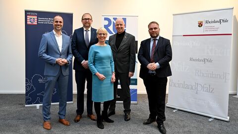 Zusammenarbeit Rhein-Main-Universitäten  Vier Männer und eine Frau lachen in die Kamera. Hinter Ihnen stehen Plakate mit den beiden Logos vom hessischen und rheinland-pfälzischen Wissenschaftsministerium.