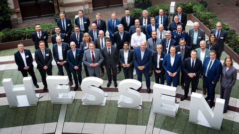 Rüstungsgipfel in Wiesbaden 2025 Über 30 Personen stehen in 3 Reihen nebeneinander in einem Innenhof und schauen nach oben in die Kamera. Vor ihnen sind 1 Meter hohe Buchstaben platziert, die das Wort "Hessen" ergeben.