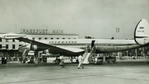 Schwarz-weiß-Aufnahme von einem Flugzeug auf dem Rollfeld. Im Hintergrund ragt ein Gebäude mit "Frankfurt Main" hervor. 