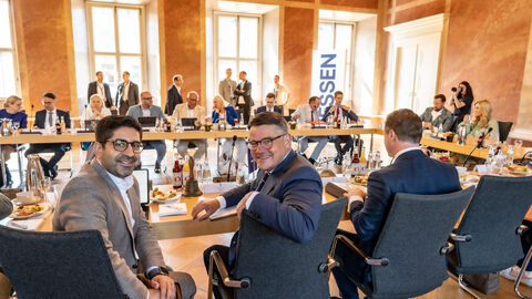 Zwei Männer im Vordergrund blicken lächelnd in die Kamera während einer Konferenz in einem historischen Saal mit einer u-förmigen Tischreihe.
