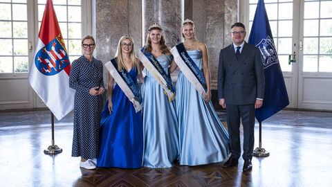 Herr Rhein und Frau Raab-Rhein stehen gemeinsam mit drei jungen Frauen in schicken Kleidern nebeneinander. Im Hintergrund stehen Flaggen.