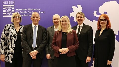 Gesundheitsministerin Stolz und weitere Personen stehen zum Gruppenfoto vor einem Aufsteller des Ministeriums.