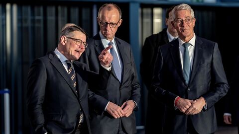 Drei Männer stehen nebeneinander. Der linke Mann zeigt mit dem Finger auf etwas außerhalb des Bildes.